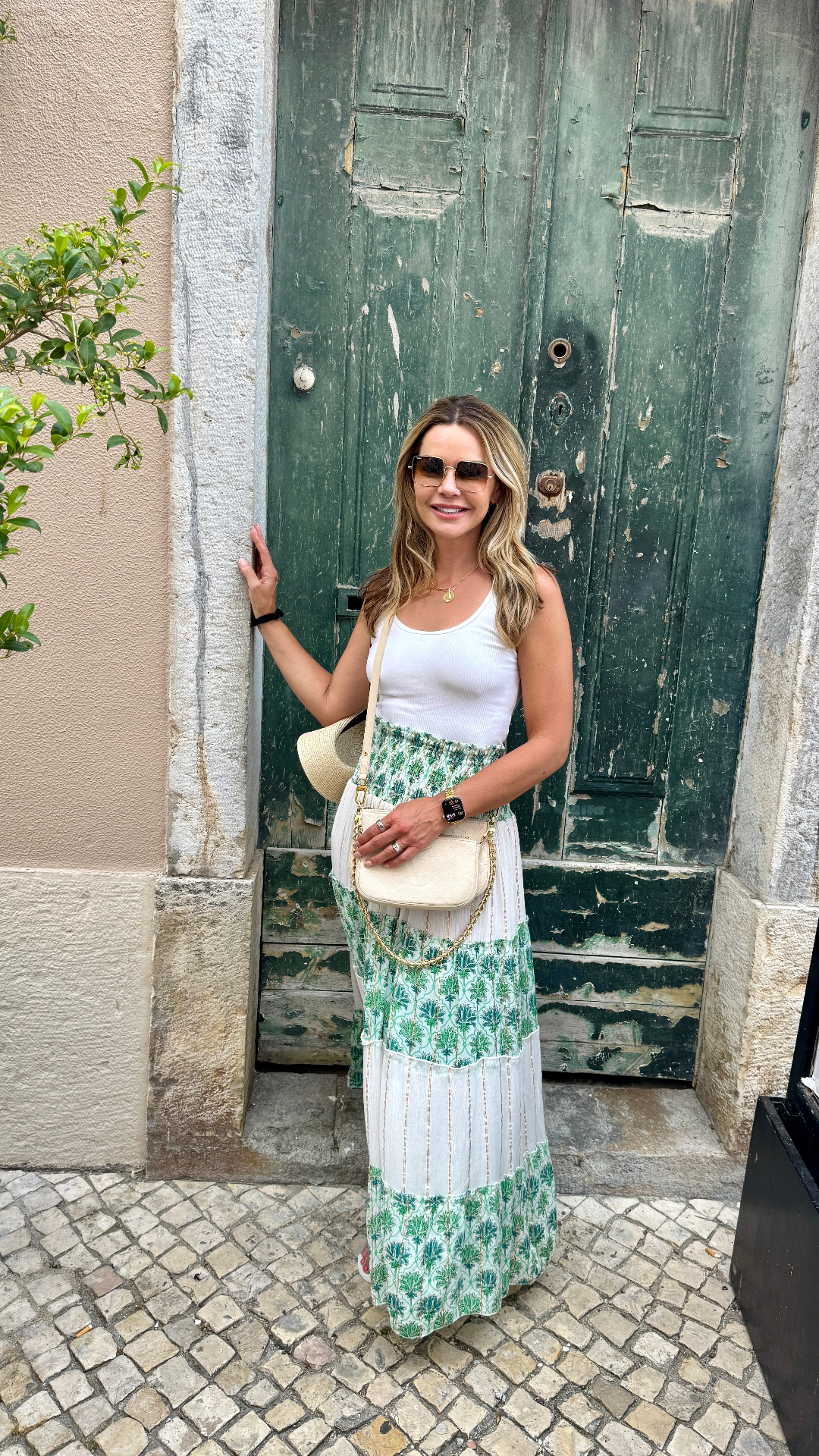 A woman standing in front of a weathered green door, wearing a white tank top and a long green and white patterned skirt with a straw hat and cream crossbody bag.