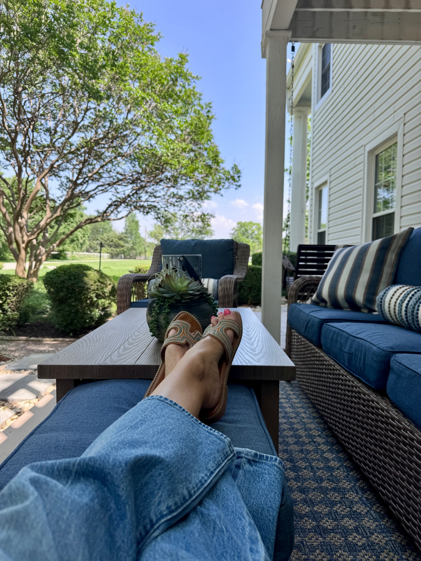 A woman's feet in sandals resting on a porch coffee table, with a sunny backyard and comfortable outdoor furniture in the background.